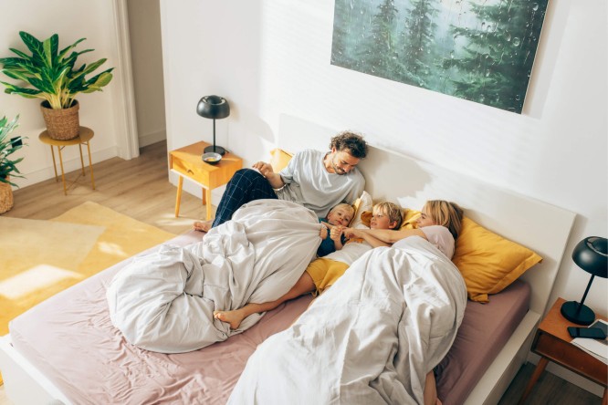 Eine vierköpfige Familie – zwei Erwachsene und zwei Kinder – entspannt sich gemeinsam auf einem Bett in einem hellen, modernen Schlafzimmer. Warme Farbtöne, natürliches Licht und Zimmerpflanzen schaffen eine gemütliche und einladende Atmosphäre. Ein Wandbild mit Waldmotiven verleiht dem Raum einen naturnahen Touch.