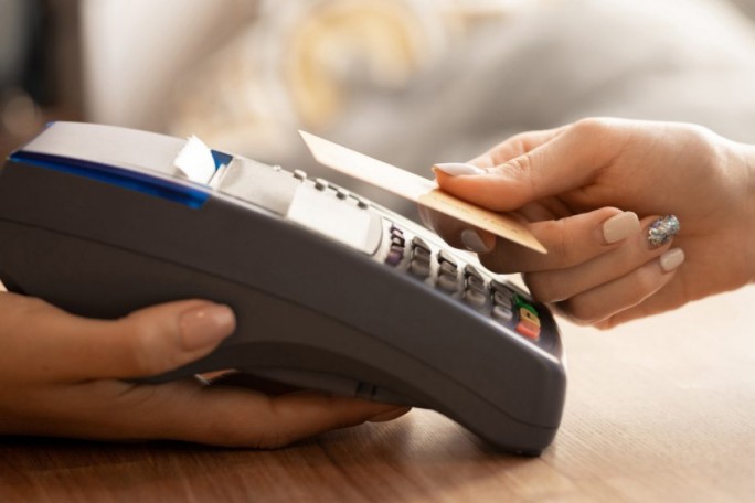 A close-up image of a person holding a card near a payment terminal for contactless transaction. The setting appears to be indoors, with a wooden surface and soft lighting. The cardholder's hand is adorned with a decorative nail design, adding a touch of elegance to the scene.
