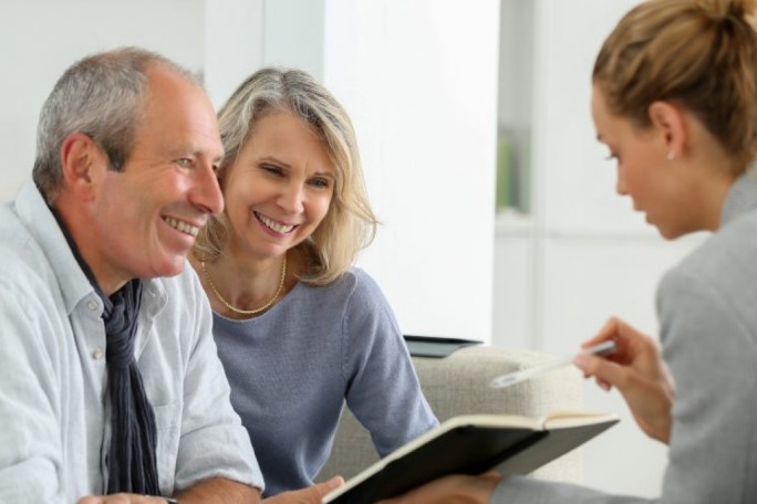 A professional meeting takes place in a bright, modern office environment. The focus is on a person holding a pen and notebook, engaging with two individuals seated on a couch. The setting suggests a formal discussion or consultation.