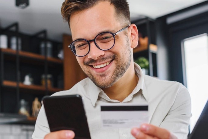 A person is seen holding a smartphone and a credit card in a modern indoor setting. The background features shelves with books and decorative items, creating a contemporary atmosphere. The individual appears to be engaged in an online transaction or payment activity.