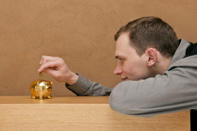 A golden piggy bank is placed on a wooden surface, with a man inserting a coin into it. The setting features a neutral brown wall in the background, creating a warm and minimalistic atmosphere. The piggy bank is shiny and reflective, emphasizing its metallic finish.