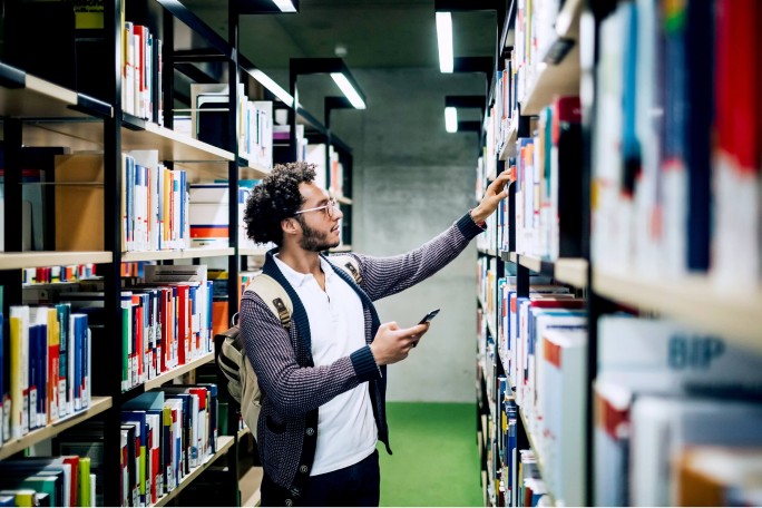 Ein junger Mann mit Rucksack stöbert in einer hellen, modernen Bibliothek. In der einen Hand hält er sein Smartphone, während er nach einem Buch im Regal greift. Die Bibliothek besticht durch ordentliche Regale voller bunter Bücher und einen grünen Teppichboden. Die Atmosphäre ist ruhig und konzentriert – ideal zum Lernen oder Recherchieren.