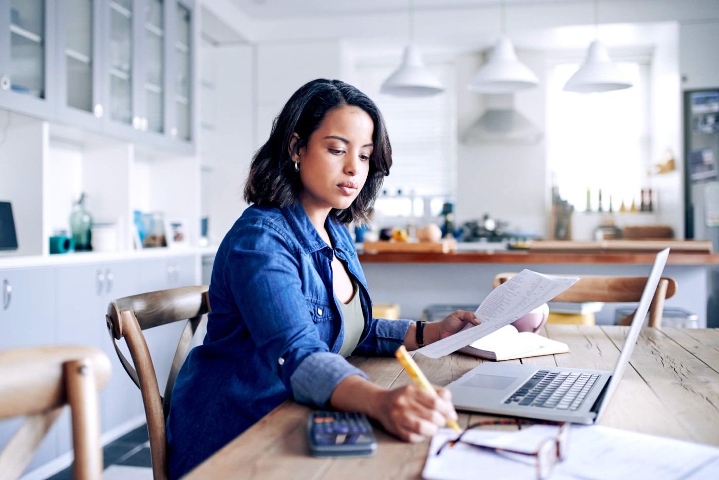 Eine Frau sitzt an einem Holztisch in einer modernen Küche und arbeitet an ihrem Laptop, während sie Dokumente durchsieht. Sie hält einen Stift in der Hand und hat einen Taschenrechner und ein Notizbuch in der Nähe, was auf finanzielle oder administrative Aufgaben hindeutet. Die helle und luftige Küche ist mit Pendelleuchten und übersichtlichen Schränken ausgestattet.