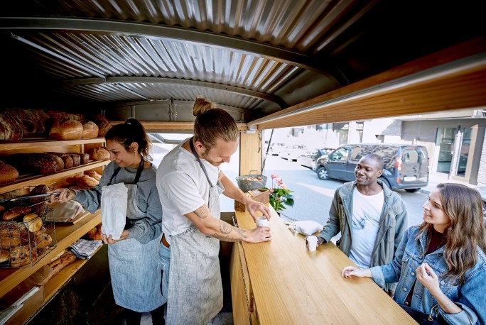 Ein mobiler Bäckereistand mit Regalen voller frischem, handwerklich hergestelltem Brot und Gebäck. Zwei Mitarbeiter in Schürzen bedienen Kunden an einem Holztresen im urbanen Umfeld. Die Szene vermittelt dank des natürlichen Lichts eine ungezwungene und freundliche Atmosphäre.