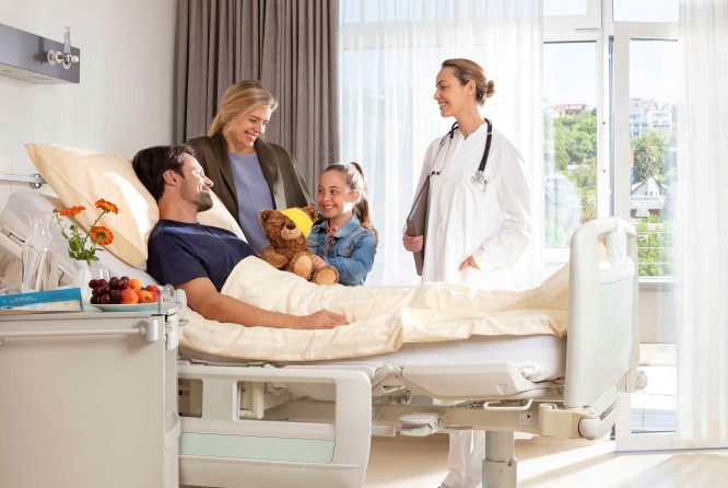 unge Familie besucht den Vater im Krankenhauszimmer, während eine Ärztin lächelnd zusieht.